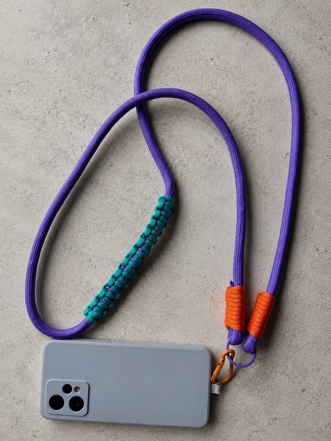 Fioletowo-turkusowy phone strap crossbody Vaporwave Sunset z pomarańczowymi detalami ułożony obok telefonu